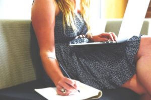 woman seated withlaptop on lap, writing on a notebook